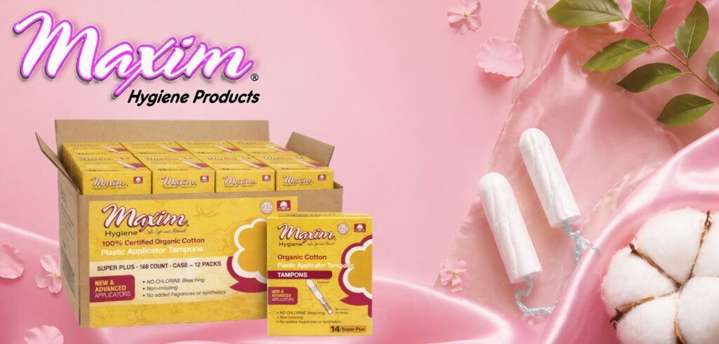 Maxim Hygiene organic cotton plastic applicator tampons displayed with retail and bulk packaging on a pink satin background with cotton flowers and green leaves.